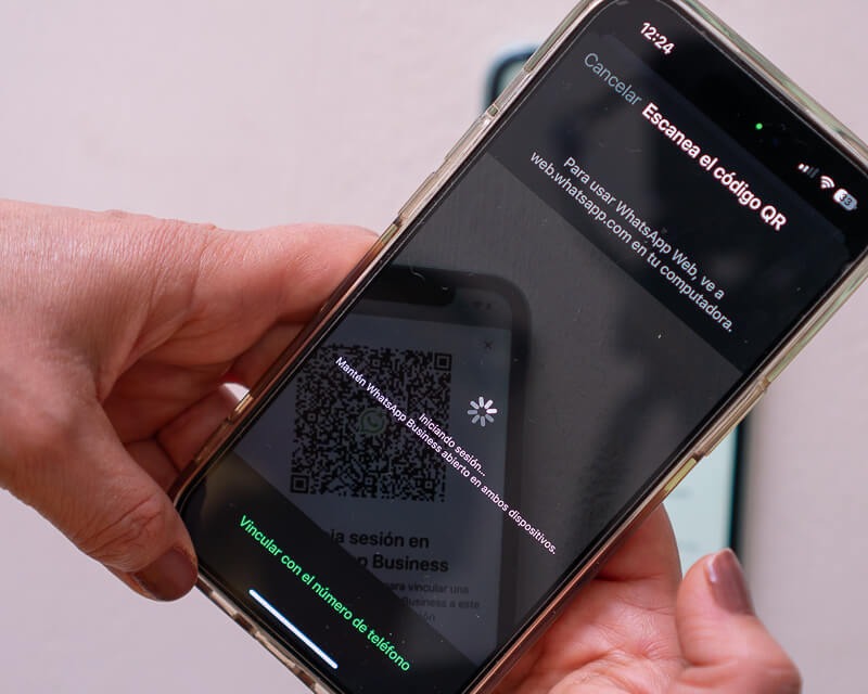 Mano sosteniendo iPhone mostrando pantalla de WhatsApp Web con código QR grande para escanear con instrucciones en texto
"Para usar WhatsApp Web, abre web.whatsapp.com en tu navegador" y opciones "Vincular con un número de teléfono" en verde. La pantalla muestra el flujo de inicio de sesión donde el usuario debe escanear el código QR con otro dispositivo para acceder a WhatsApp.